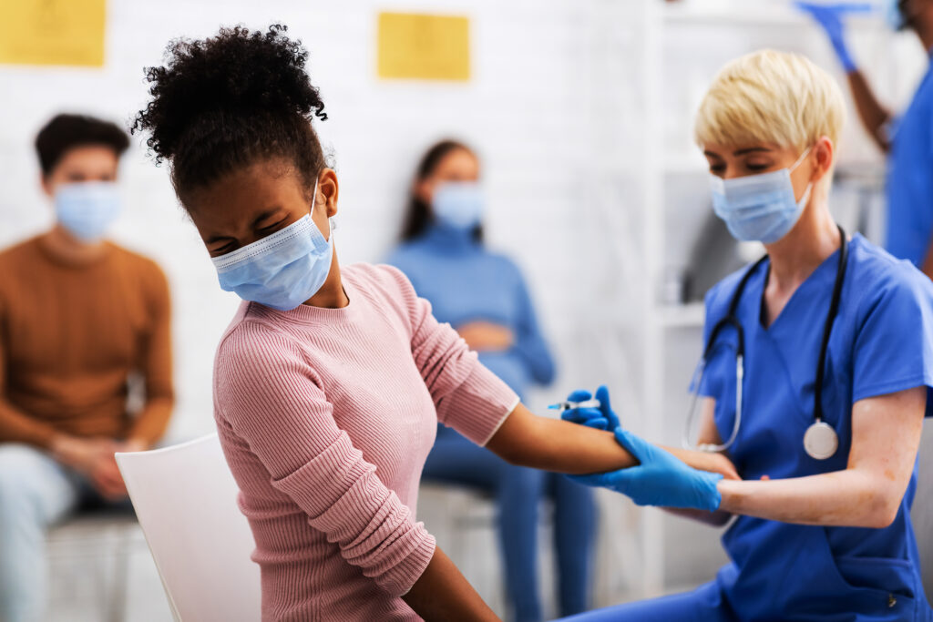 冠状病毒疫苗接种。 黑人青少年女孩接受covid-19疫苗注射在手臂与护士在医院的病房里坐着。 电晕病毒的预防，治疗和药物治疗的概念。 选择性焦点
