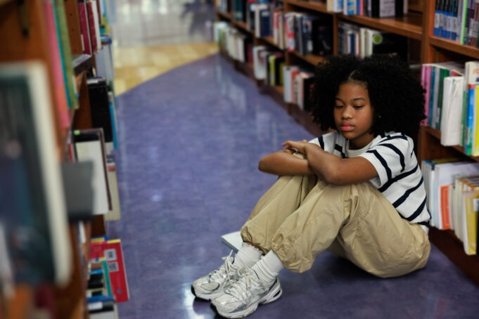 An overwhelmed and sad young African American schoolgirl sitting on the floor between bookshelves in a library, hiding her face in her arms.