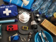 Are you and your family prepared? A collection of emergency supplies including a first aid kit, face mask, flashlight, water bottles, canned food, crackers, a radio, and utensils arranged on a surface. Description generated by AI