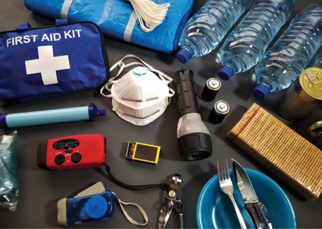 Are you and your family prepared? A collection of emergency supplies including a first aid kit, face mask, flashlight, water bottles, canned food, crackers, a radio, and utensils arranged on a surface. Description generated by AI