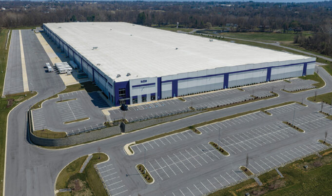 A large industrial warehouse with a white roof and blue accents is surrounded by empty parking lots and roads in a rural area. Description generated by AI
