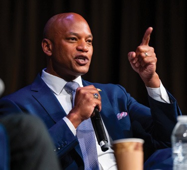 A person in a blue suit and tie is speaking into a microphone while gesturing with one hand during an event. Description generated by AI
