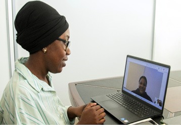 A person wearing glasses and a headwrap is sitting at a table and engaging in a video call on a laptop with another person visible on the screen. Description generated by AI