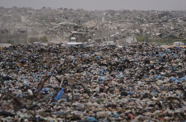 The valley of decision A vast expanse of garbage and debris stretches across the foreground with a densely packed area of heavily damaged and ruined buildings in the background under a gray sky. Description generated by AI
