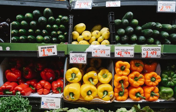 Green squash, yellow squash, cucumbers, red and yellow bell peppers, and other vegetables are displayed on shelves with price tags in a grocery store.

Description generated by AI