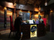 Africa’s mother tongues are finding new life as youth connect with roots and relatives Three adults wearing traditional headwraps are seated around a decorated wooden table in a warmly lit room with patterned walls and a chalkboard menu behind them. Description generated by AI