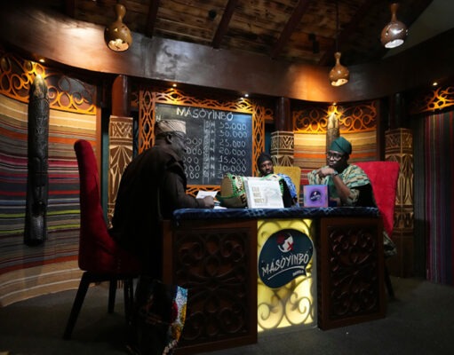 Africa’s mother tongues are finding new life as youth connect with roots and relatives Three adults wearing traditional headwraps are seated around a decorated wooden table in a warmly lit room with patterned walls and a chalkboard menu behind them. Description generated by AI