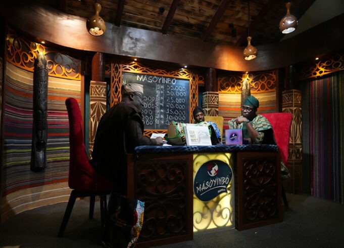 Three adults wearing traditional headwraps are seated around a decorated wooden table in a warmly lit room with patterned walls and a chalkboard menu behind them. Description generated by AI