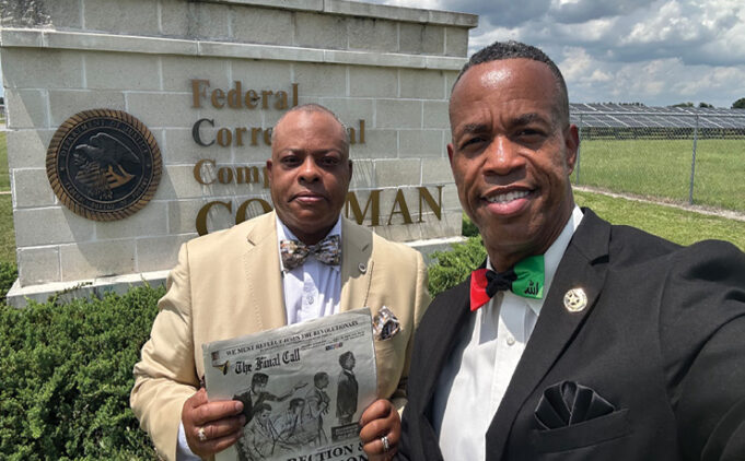Two adults dressed in formal attire stand outdoors in front of a sign for a federal correctional complex under a partly cloudy sky. Description generated by AI