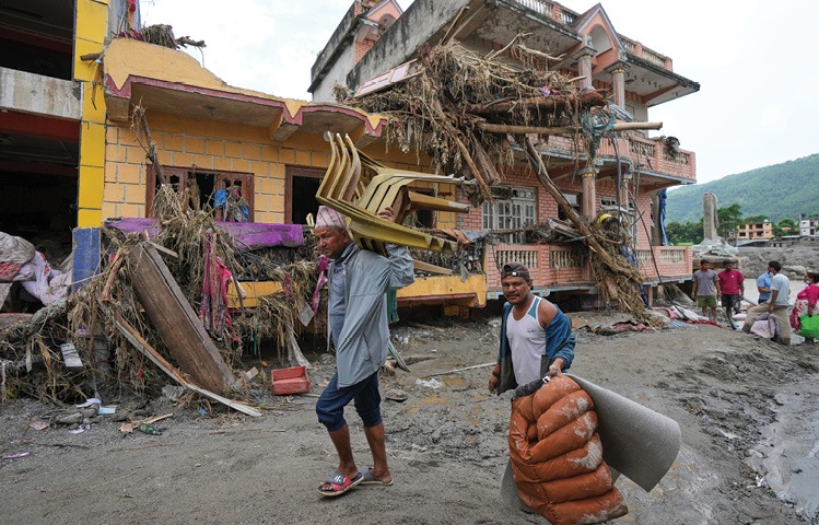 Two people carry chairs and bags through a muddy area in front of damaged buildings covered with debris from a natural disaster. Description generated by AI